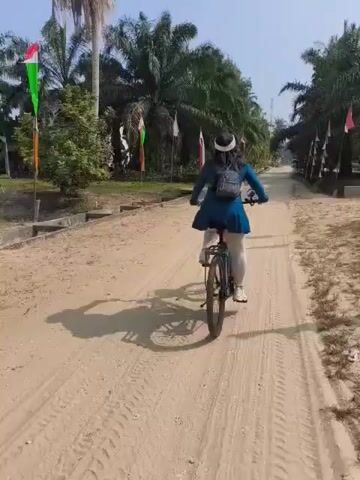 A person is riding a bicycle on a dirt road. The person is wearing a blue tunic, white leggings, and a black headscarf with a white band. They are also wearing a small gray backpack. The bicycle is black and has a rack on the back. The road is sandy and has tire tracks. There are palm trees on both sides of the road, and some flags are visible in the background. The sky is clear and the lighting suggests it is daytime. The camera is following the cyclist from behind.