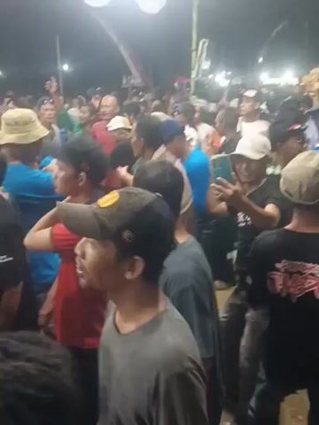 The video shows a crowded outdoor gathering at night, illuminated by bright overhead lights. Many people are visible, most of them men, dressed in casual attire including t-shirts, hats, and some in what appear to be uniforms. Several individuals are holding up their mobile phones, presumably to record the event. The atmosphere appears energetic, with some people raising their hands or gesturing. In the foreground, a man wearing a gray t-shirt and a black baseball cap with a logo is looking upwards and to his right, with his mouth open. To his left, another man in a red t-shirt and a camouflage baseball cap is visible. Further back, a person in a blue t-shirt is holding a phone and appears to be recording. The ground is uneven and appears to be dirt or sand. The overall impression is of a lively public event or celebration.
