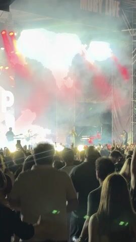 The video shows a crowd of people at an outdoor concert. The stage is lit with red and white lights, and there is smoke or fog in the air. The band or performer is visible on stage, and the crowd is enthusiastically cheering and raising their hands. The video also shows the audience from behind, with many people holding up their phones to record the event. The overall atmosphere is one of excitement and enjoyment.