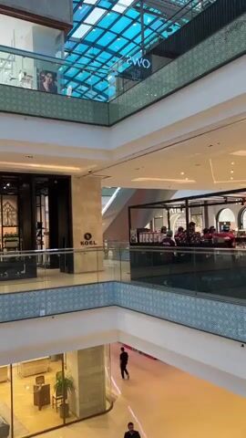 The video pans across the interior of a multi-level shopping mall. The architecture features modern design with glass railings and spacious floors. On the upper levels, storefronts are visible, including one labeled "OWNS" and another with "BY HEMANI." Further down, a store named "NUREH" displays clothing. People are seen walking on different levels of the mall, some browsing in shops and others walking through the common areas. Escalators and elevators are also present, indicating the vertical connectivity of the mall. The lighting appears to be a mix of natural light filtering from above and artificial mall lighting. The overall atmosphere is that of a typical, busy shopping environment.