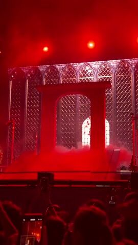 The video captures a concert stage set bathed in intense red lighting. The primary subject is a large, monolithic, red, arch-shaped structure that dominates the center of the stage. Behind it, a backdrop resembling a gothic window with intricate latticework adds to the dramatic atmosphere. Smoke or fog emanates from the stage floor, particularly around the base of the arch, creating an ethereal and mystical effect.

The scene suggests a live performance, likely a concert or a theatrical show, given the stage setup and the presence of microphones on stands at the sides. The red lighting creates a powerful and possibly intense mood, amplified by the smoke. The backdrop with its window-like pattern could be a projection or a physical set piece designed to evoke a specific aesthetic, perhaps gothic or ancient.

While the faces of the performers are not clearly visible, the focus is on the stage design and the overwhelming red ambiance. The presence of an audience is indicated by the silhouettes of people and hands holding up phones at the bottom of the frame, suggesting they are actively recording or photographing the event. The emotion conveyed is one of spectacle and anticipation, amplified by the dramatic lighting and staging.

There is no discernible text visible in the video. The location context provided, Segrate, Italy, helps to place this event in a specific geographical area, but there are no visual cues within the video itself to confirm this location. The time of day is not evident, but the indoor setting and the lighting suggest it could be nighttime or a deliberately darkened venue. The weather is irrelevant as the scene is indoors.