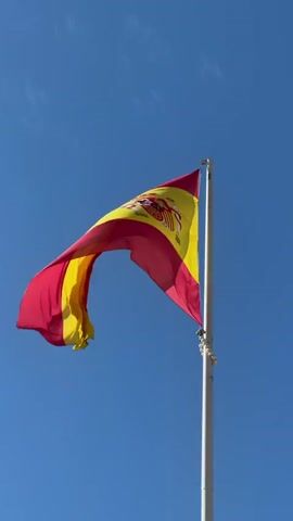 A Spanish flag flies in the wind against a clear blue sky. The flag is attached to a white flagpole. The flag consists of three horizontal stripes: red, yellow, and red. The yellow stripe is wider than the red stripes and features the Spanish coat of arms in the center. The flag is blowing to the left, causing it to ripple and wave.