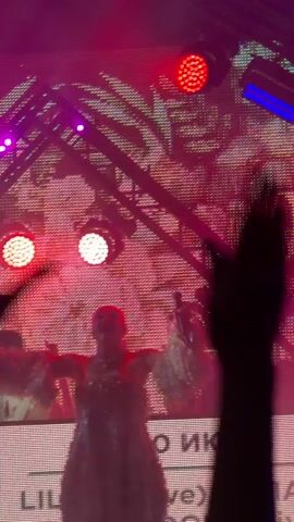 A concert stage with bright pink and blue lights. A person is on stage, holding a microphone and wearing a sparkly outfit with fluffy accents. Another person, likely a DJ, is behind a table with lights. The audience is visible in the foreground, with hands raised in the air. The background features a large screen with a geometric pattern and lights. Text is visible on a banner at the bottom of the screen, reading "LIL" and some Cyrillic characters. The overall atmosphere is energetic and festive.
