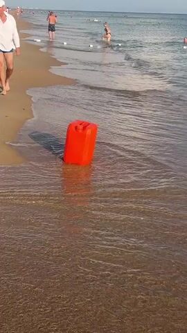 The video begins with a close-up of a bright orange plastic jerrycan partially submerged in the shallow water at the edge of a sandy beach. The waves gently lap around it. The camera then pans out to reveal the wider beach scene. In the distance, several people are visible in the water, some swimming and others near buoys marking a swimming area. The sea is calm with small waves breaking on the shore. Further out, a yellow inflatable raft with several people on it is being towed by someone in the water. The person towing the raft is holding a rope. The sky is clear and blue. The scene appears to be a typical summer day at a beach. The accompanying audio states that this is a reserve of gasoline due to a problem with it.