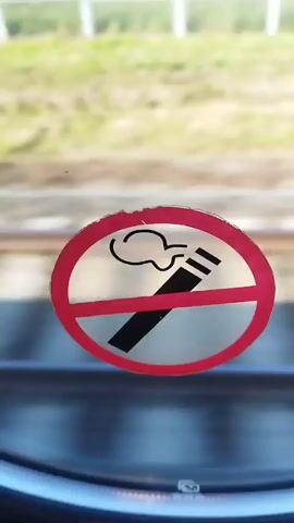 The video begins with a close-up of a round, red-bordered sticker on a window, depicting a black cigarette with smoke and a red diagonal line through it, indicating "no smoking." The background outside the window is blurred, showing green foliage and a white fence. The camera then pans to the right, showing the same sticker as the view outside the window changes to a blurred landscape of green grass, trees, and a blue sky, suggesting movement. The train tracks are visible in the foreground.