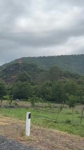 A video captured from a moving vehicle shows a church on a hilltop surrounded by trees. The sky is overcast, with gray clouds dominating the frame. The foreground features a gravel path and a grassy area with a few young trees. In the middle ground, dense green trees create a lush backdrop. The church, with its red-tiled roof, is situated on a rocky outcrop on the distant hillside. The camera pans slightly, revealing more of the mountainous terrain and the vegetation. The overall impression is of a serene, natural landscape with a prominent religious structure.