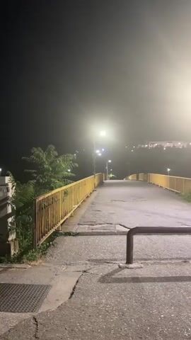 The video shows an empty pedestrian bridge at night. The bridge is illuminated by streetlights, which cast long shadows. The sky is dark and hazy, suggesting fog or smog. The bridge is bordered by a yellow railing. There is some vegetation visible on either side of the bridge. The path on the bridge is paved with asphalt, and there are some cracks and unevenness in the surface. In the distance, there are more lights, possibly from buildings or other streetlights. A dark metal railing is in the foreground, positioned horizontally across the path.