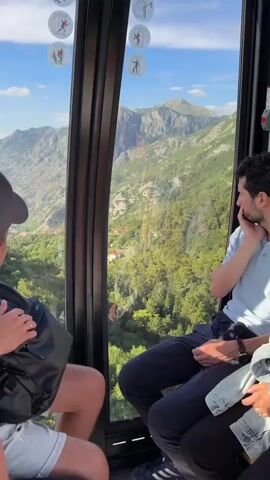 The video shows the interior of a gondola car moving uphill with several people inside. Through the large windows, a vast mountainous landscape with a town nestled in a bay is visible. Sunlight illuminates the scene, casting shadows on the interior. The gondola car has a dark metal frame and floor. People are seated and looking out the windows. One person in the foreground wears a dark baseball cap and a black backpack. Another person, in the middle right, is wearing a light blue shirt and dark pants. A third person, on the far right, wears a denim jacket. Small circular stickers with icons of people in motion are affixed to the top of the windows. The view outside includes lush green trees covering the slopes, winding roads, red-roofed buildings, and a body of water. The sky is blue with scattered clouds, and the distant mountains are covered in lighter green vegetation.