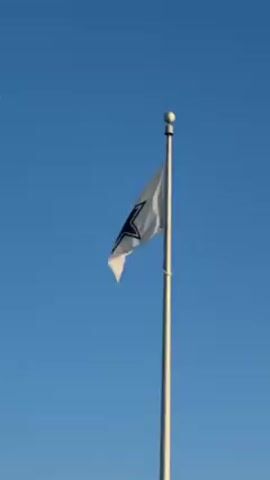The video shows a Dallas Cowboys flag waving in the wind against a clear blue sky. The flag is white with a navy blue star in the center. It is attached to a tall, silver flagpole topped with a spherical finial. The camera angle is low, looking up at the flag.