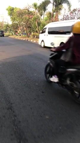 The video shows a street scene with several vehicles. In the foreground, a white car is driving away from the camera. To the left of the white car, a motorcycle with a rider wearing a red jacket and yellow helmet is also moving away. Further in the background, a white van is visible. The environment appears to be a road with trees and foliage on the sides. The lighting suggests it is daytime, possibly late afternoon. The camera appears to be mounted on a moving vehicle, as indicated by the motion blur.