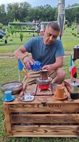 A man with short brown hair, wearing a blue t-shirt and brown shorts, kneels in front of a wooden crate that serves as a table. He is pouring water from a clear blue bottle into a dark metal pot, which sits on a red camping stove. Two small flags, one blue and yellow and the other red, white, and green, are planted next to the pot. On the crate, there is a silver bowl, a blue and white spotted mug, a small glass with a silver saucer, an orange mug, a silver mocha pot, and another glass with a saucer. In the background, there is a grassy field with trees and what appears to be a display of vintage aircraft, including a helicopter and a plane.