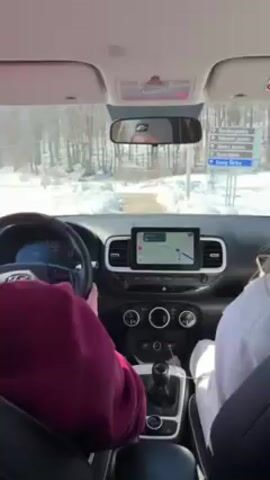 From inside a car, a driver in a burgundy shirt steers down a snowy road past trees. A passenger in a white hoodie holds a phone and touches the screen. The car's dashboard is visible with a navigation display.