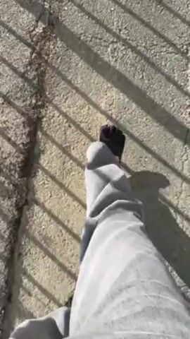 The video shows a person walking on a cracked concrete path with shadows of a metal railing overhead. They then move to a viewpoint protected by a metal railing. The view overlooks a lake surrounded by mountains, with the ocean visible in the distance under a sunny sky.