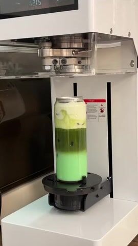 A can-shaped glass filled with a three-layered green and white beverage sits on a black turntable inside a white machine. The bottom layer is a pale green, the middle layer is a darker green with dark spheres inside, and the top layer is a white, frothy topping. The machine has a silver, block-like component above the glass. A red warning sticker is visible on the side of the machine. The lighting is bright and the background is out of focus.