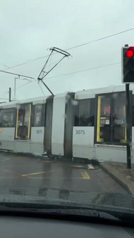 A Metrolink tram, numbered 3075, is stopped at a traffic light. The tram is white with yellow accents and has the Metrolink logo visible. Inside the tram, passengers can be seen through the windows. The tram is on a wet road with yellow lane markings, suggesting it is raining or has recently rained. A traffic light above and to the right of the tram is red, indicating that traffic should stop. The view is from inside a vehicle, with a windshield wiper visible in the foreground. In the background, other cars are stopped behind the tram, and buildings and streetlights line the road. The sky is overcast and gray.