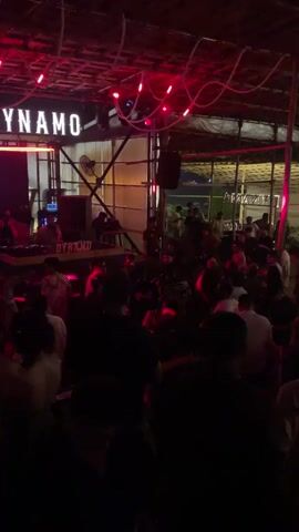 The video depicts a crowded indoor event, possibly a nightclub or bar, with a significant number of people dancing and socializing. The environment is dimly lit, with vibrant red neon lights creating a festive atmosphere. Above the crowd, a sign displays the word "DYNAMO" in illuminated letters, indicating the name of the venue or an event. A DJ booth is visible on a raised platform to the left, with individuals operating audio equipment. The ceiling appears to be made of bamboo or thatched material, adorned with string lights. The overall scene conveys a sense of energetic nightlife and entertainment.