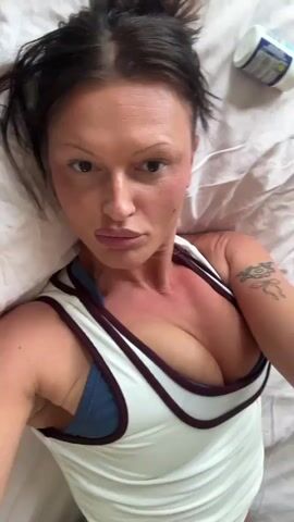 This media file captures a close-up, low-angle shot of a woman lying down on a white surface, likely a bed with a white pillow. She has dark hair, light skin with noticeable redness on her cheeks and chest, suggesting potential sunburn or makeup. She is wearing a white ribbed tank top with dark trim.

The woman appears to be looking directly at the camera, with varying expressions. In some frames, her lips are slightly parted, and in others, she has a small white object in her mouth, possibly food or a pill. The lighting suggests it is daytime, but there are no clear external environmental cues to confirm time of day or weather. The setting is indoors and intimate, focusing solely on the woman and her immediate surroundings. There is no visible text in the media file. The activity appears to be a casual moment, perhaps a selfie or a personal video recording.