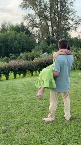 A man carrying a woman in his arms in a grassy outdoor setting. The man is wearing a light blue t-shirt and light-colored pants. The woman is wearing a bright green dress. They are both looking at each other and appear to be smiling or kissing. In the background, there are trees and a row of purple flowers. The lighting suggests it is either early morning or late afternoon.