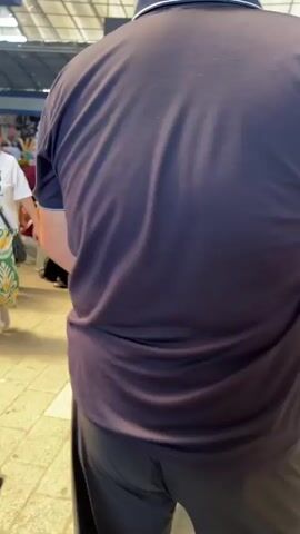 A man in a dark t-shirt and black pants walks towards the camera. In the background, a busy marketplace is visible with stalls and people. A woman in an orange shirt bends down near a trash can. An ice cream cart with pictures of ice cream is set up. A woman wearing a red hijab pushes a baby stroller. Another woman in a red shirt and hijab is visible. People are walking around the market.