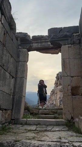 The video features a woman standing within an ancient stone archway, framed by eroded stone walls. She has long dark hair, wears glasses, a dark jacket over a dark skirt or dress with a white and black stripe, and white sneakers. A colorful scarf or garment is draped over her arm.

The woman is posing for the camera. Initially, she raises her hand to her mouth, making a gesture, and then lowers it, shifting her gaze. Her expression appears relaxed and content.

The archway is made of large, weathered grey stones, suggesting it's part of a historical ruin. The ground inside the archway is also stone, with small plants and weeds growing through the cracks. Beyond the archway, a expansive valley is visible, with distant mountains under an overcast or cloudy sky, indicating either early morning or late afternoon light. The scene conveys a sense of historical exploration and appreciation for natural beauty, likely at an archaeological site in Finike, Turkey. No visible text is present.