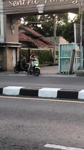 The video shows a street scene with several motorcycles. In the foreground, a curb with black and white markings is visible. Behind the curb, the asphalt road is seen. In the middle ground, a green motorcycle with two people on it, a driver and a passenger, is turning. To the left, another motorcycle with one person is moving away. Further to the right, a third motorcycle is partially visible, its front wheel spinning as it moves. In the background, there are buildings, vegetation, and a blue gate. The overall lighting suggests it is either early morning or late afternoon.