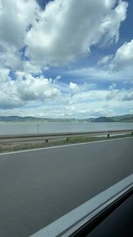 The video shows a view from inside a vehicle, looking out a window towards a wide body of water bordered by hills under a partly cloudy sky. A road with a guardrail runs parallel to the water. The clouds are white and puffy, with patches of blue sky visible between them. The water appears calm, and the hills in the distance are covered in green vegetation. The road surface is asphalt, and the guardrail is metallic. The time stamp indicates the video is from the beginning of a recording.