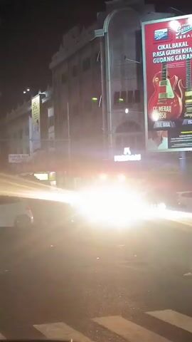 At night, a street scene unfolds with a prominent clock tower on the left and buildings lining the right side of the street. A white van, identified by markings including "BUSLAS" and "M. DAUD, SE" on its side, is stationary in the middle of the road, emitting a bright light that illuminates the surroundings. The van appears to be engaged in some activity that involves spraying a substance, possibly water or a cleaning agent, across the road. Several cars and motorcycles are visible on the road, some passing by, others stopped. Large billboards are erected on the buildings, one prominently displaying a "76 APEL ROYAL" advertisement. The street is paved, with visible white road markings. The overall atmosphere suggests an urban environment at night, with artificial lighting from street lamps, vehicle headlights, and billboards illuminating the scene.