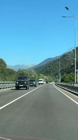 The video shows a POV shot from a car driving on a highway. The road is asphalt with white lane markings. There are other vehicles on the road, including cars and vans. The highway is elevated and bordered by guardrails. On either side of the highway are lush green hills and mountains. The sky is clear and blue. There are streetlights along the side of the road. The overall scene is one of travel through a scenic mountainous landscape.