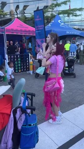 A group of people are gathered outdoors in what appears to be a street festival or community event. In the foreground, a large grey stroller with a blue bag and a purple bag is visible. To the right of the stroller, two young women are standing, one with dark hair and a pink top and skirt, and the other with lighter hair and a purple top. Behind them, a large blue tent is set up with the Prospera logo. To the left of the stroller, a large connect four game is set up, with several children playing or interacting with it. Two tents with pink and blue branding are also visible in the background, as well as several adults and children milling around. The environment appears to be urban, with a modern building with reflective windows in the background. The lighting suggests it is daytime.