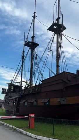 A video taken outdoors features a close-up view of a wooden sailing ship's bow. The ship has multiple masts and rigging, with dark wooden planks forming its hull and lighter wood accents. Small, rectangular windows are visible along the side. The ship is on display on land, with a green lawn in the foreground and a metal fence. Behind the ship, there are a few small buildings, a red post, and a white striped barrier. The sky above is blue with scattered white clouds. The camera pans slightly to the right, revealing more of the ship's structure.