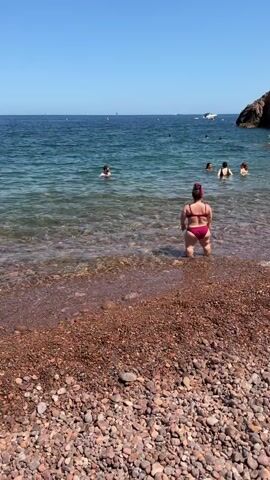 A person wearing a red bikini stands in shallow water at a beach. The person is facing away from the camera. The water is clear and reflects sunlight. The beach is covered in small, reddish-brown pebbles. In the background, other people are swimming and enjoying the water. The sky is clear and blue. The scene is sunny and appears to be a warm day.