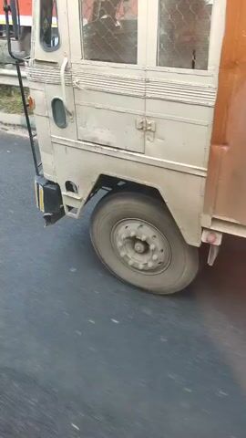 The video shows the front right quarter of a light-colored truck as it drives on a paved road. The truck appears to be utilitarian, possibly a cargo vehicle or a specialized service vehicle, with a cage-like structure visible through the windows. The road surface is asphalt, and there is some debris visible on the road. The truck's tire is in motion, indicating the vehicle is driving. The lighting suggests daytime.