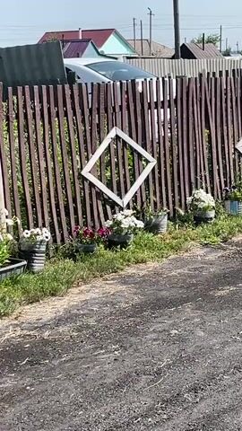 The video shows a wooden fence with vertical planks. There are several white diamond shapes attached to the fence. In front of the fence, there are flower pots with white and pink flowers. There is also some green grass and a dirt path in the foreground. In the background, there are houses and trees. The sky is blue and there are some clouds. The video is filmed from a slight downward angle, as if the camera is being held by someone walking or driving past the fence.
