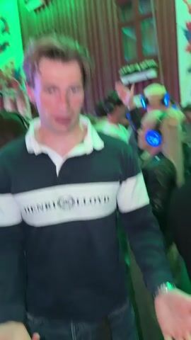 A man in a navy and white Henri Lloyd rugby shirt with a white collar and a white stripe across the chest is standing in the foreground, facing towards the viewer. He has light brown hair and is speaking, with his hands gesturing outwards and upwards. He is wearing a ring on his left hand. In the background, other people are visible wearing headphones. Some are dancing or moving to music. The environment appears to be indoors, possibly a venue or party, with wooden paneling on the walls and green lighting. A large banner with images and text is visible on the left side of the frame. A woman in a black leather jacket and a white top is in the middle ground, also wearing headphones and raising her hand. The overall atmosphere seems to be a silent disco or a similar event where participants listen to audio through headphones.