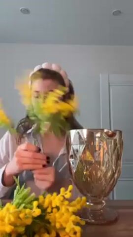 Here's a description of the video:

A woman with fair skin, wearing a white button-down pajama top with red piping and a white headband, is arranging yellow flowers in a gold-colored, geometric-patterned glass vase. She is seated at a light-colored wooden table. Some of the yellow flowers are already sitting on the table. She holds a bouquet of yellow flowers with green leaves, examining them, and then places them into the vase. The background is a white wall. Two ceiling lights are visible. A white door frame is also visible behind her.