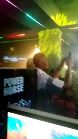 The video shows a crowd of people in a dark, smoky room that appears to be a nightclub or event venue.  There are various colored lights overhead, including green, red, and blue, creating a party atmosphere. Some people in the crowd are holding up their phones, possibly recording or taking pictures. A person in the foreground is partially obscured by a computer monitor and a black object with the words "POWER HORSE" printed on it. The overall impression is a lively and energetic social gathering.