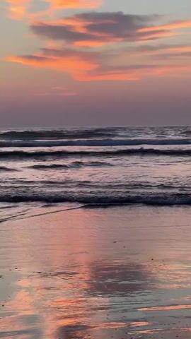This is a vertical video of a beach at sunset. The sky is filled with pink and orange clouds. The ocean has several waves rolling in. The wet sand reflects the colors of the sky. There are no people or objects visible in the video. The overall mood is peaceful and serene.