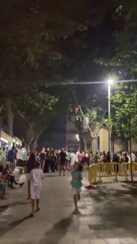 The video shows a nighttime street scene with many people gathered. Trees with foliage overhang the street, and there are streetlights illuminating the area. Some people are sitting on the side of the street, while others are standing and walking. In the foreground, two children are walking down the street. The crowd is dense further down the street, with some individuals being carried on others' shoulders. Buildings line the street, and some windows appear to be lit. There are also some yellow barriers visible. The overall atmosphere seems to be one of a lively street event or festival.