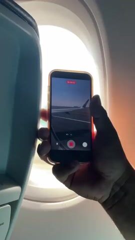 A person is holding a smartphone to record video through an airplane window. The phone screen shows a view of the airplane wing and runway. The window shows the wing of the plane is green and has the letters "Viva" on it, as well as a view of the airport outside.