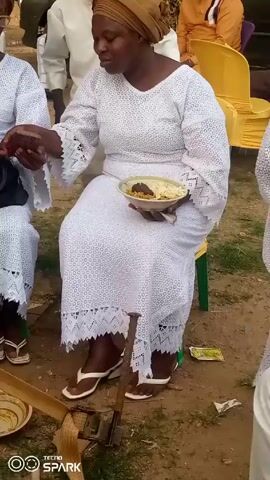 A seated woman in a white lace dress and brown headwrap holds a plate of food and looks to her left. Other people wearing similar white outfits are seated around her. Some are wearing white clothes. The setting appears to be outdoors on a dirt surface, with yellow chairs visible in the background. A TECNO SPARK watermark is visible in the lower left corner.