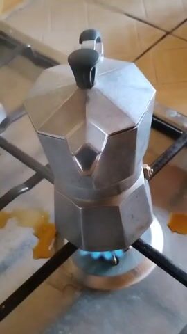 A silver moka pot sits on a gas stove with a blue flame underneath. There are some brown spills on the stovetop around the burner. The stovetop has a stainless steel surface with black grates. The background shows beige-colored tile walls. Steam rises from the moka pot as the coffee brews.