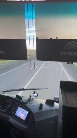 The video shows the interior of a bus, looking out through the windshield. The scene is a road in Kazakhstan, specifically near Шешенқара, during the daytime. The weather appears to be clear with a bright sky.

The primary focus is the bus driver's perspective. We see the dashboard of the bus, including the steering wheel, various buttons and controls, and a digital display screen. On the screen, there is an image of a couple. A driver, a man with light-colored hair and wearing a light-colored shirt and beige pants, is visible in the driver's seat, his hands on the steering wheel. The bus is in motion, driving on a paved road with a white line marking the lanes. In the distance, other vehicles can be seen on the road.

There is text visible on the interior of the bus, above the windshield. On the right side, the word "YUTONG" is printed in a logo. On the left side, another logo or text is partially visible. There are also some signs in Russian above the windshield, including one that appears to say "НЕ КУРИТЕ" (Do not smoke).

The overall impression is a view from the front of a moving bus on a highway in Kazakhstan. The time of day is likely late afternoon or early evening, given the quality of the light. There are no strong emotional cues, but the scene conveys a sense of travel and movement.