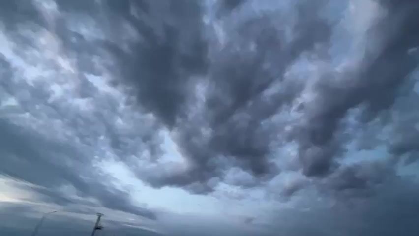 The video shows a wide shot of a cloudy sky. The clouds are dark gray and appear to be moving. The sky is a light blue color in between the clouds. There is a faint hint of a pole or structure on the left side of the frame, possibly a utility pole. The footage is shaky and appears to be handheld.