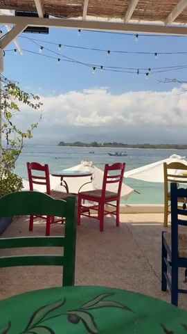 The video shows an outdoor cafe with a view of the ocean. In the foreground, there are green chairs, and in the middle ground, there is a small round table with two red chairs. Beyond the table, there are white umbrellas and lounge chairs on the beach. The ocean is a clear blue, and in the distance, there are islands and boats on the water. The sky is blue with some white clouds, and there are string lights overhead. The cafe has a thatched roof.