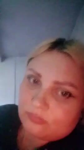 The video shows a close-up, low-angle shot of a woman with blonde hair. She has freckles on her face and is wearing makeup, including eyeliner. Her expression appears thoughtful or perhaps a little sad, with her eyes looking down and to the side. The lighting is dim and warm, suggesting it might be evening or indoors with artificial light.

The setting appears to be indoors, with a grey ceiling visible in the upper portion of the frame and a white wall or door in the background. A black garment, likely clothing, is visible on the left side of the frame.

The woman speaks in Russian, with the timestamp "00:02" overlaid on the video showing the text "Ой, у нас такая жара" which translates to "Oh, we have such heat." At timestamp "00:03", the text "00:03 наконец-то я дома приехала" appears, translating to "00:03 finally I arrived home." This indicates the woman has just returned home after being out in hot weather.

The overall mood is intimate and personal, as it is a close-up selfie-style video. The mention of heat and arriving home suggests a recent journey or a period of being outside. The location context of "Изыхские Копи, Russia" is provided but not directly visible in the video itself.