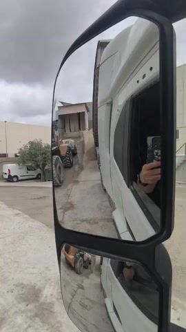 The video shows a truck's side mirror, reflecting a scene outside the vehicle. The mirror view includes the side of the white truck itself, along with a large construction vehicle (possibly a front loader) parked further down a concrete path.  A smaller, orange construction vehicle is visible in the lower portion of the mirror.  A pile of gravel or sand is located near a building with a loading area. In the background, a white van is parked near a tree. The sky is overcast.  The person inside the truck is holding a phone, presumably recording this video. The person's hand and phone are visible in the truck's window reflection within the mirror.