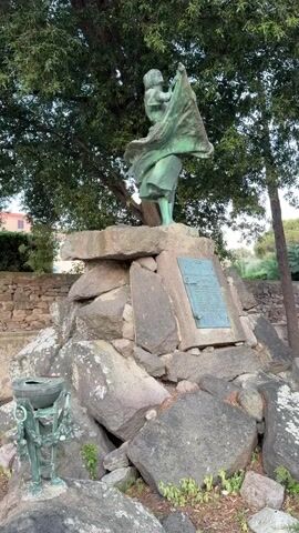 The video shows a statue of a woman holding a baby in her arms while appearing to wave a flag or cloth. The statue is made of a greenish metal, likely bronze, and stands on a base of large, rough-hewn rocks. A bronze plaque is affixed to one of the rocks on the right side of the statue. To the left of the statue, there is a smaller bronze object that appears to be a vase or urn supported by a decorative stand. The statue and its base are situated outdoors, with a large tree providing shade from above. Greenery and more rocks are visible around the base.