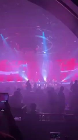 The video shows a live performance on a stage illuminated with purple and blue lights. The stage has a large screen displaying abstract graphics. On the stage, there's a group of at least nine performers, mostly women, dressed in varying outfits including leotards and short dresses. They are performing a choreographed routine, with some visible movement and gestures. In front of the stage, a crowd of people is watching the show. Some individuals are holding up phones, seemingly to record the performance. The environment suggests an entertainment venue, likely a nightclub or theater, with a lively atmosphere.