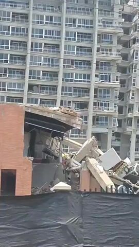 A partially collapsed building is shown. The building appears to be a high-rise with multiple stories and many windows. The lower section of the building has significant damage, with debris scattered around it. A black fence with a tarp is in the foreground, partially obscuring the view of the damage. In the background, a taller, intact section of the building stands. A police vehicle with flashing lights is visible in front of the damaged section. The sky is overcast.