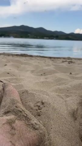 A low-angle close-up shot shows a sandy beach with a person's sand-covered foot in the foreground. In the background, a body of water stretches to the horizon, with rolling hills and a cloudy sky above. The water is calm with gentle ripples. The scene appears to be a natural outdoor environment, possibly a lake or a large pond. The lighting suggests it is daytime, possibly late afternoon or early evening due to the soft light and clouds. There is no discernible audio track.
